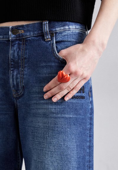 Red heart-shaped ring made of glossy material worn on fingers, featured against blue denim jeans with subtle texture and stitching details.