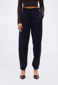 Γυναίκα που φοράει μπλε σκούρο στενά παντελόνια με ελαστικές μανσέτες, μακρυμάνικη crop μπλούζα σκούρου καφέ χρώματος και μαύρες μυτερές slingback γόβες, όρθια.