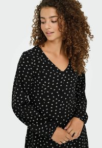 Jeune femme aux cheveux bouclés portant une robe noire à manches longues avec des motifs de nœuds blancs, regardant vers le bas avec les mains doucement jointes.