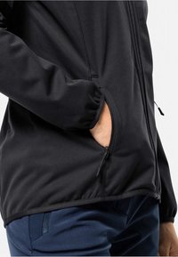 Svart softshell-jakke med en glidelåslomme, glatt tekstur og elastiske mansjetter. Sammen med marineblå bukser og en tettsittende design.