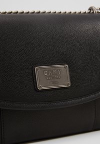Sac à main DKNY en cuir noir avec une bandoulière chaîne argentée et une plaque logo métallique rectangulaire portant l'inscription "DKNY New York EST. 1989."