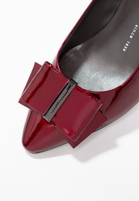 Chaussure en cuir verni bourgogne à bout pointu avec un grand nœud sur le devant, rehaussé d'un petit logo en métal. Texture lisse.
