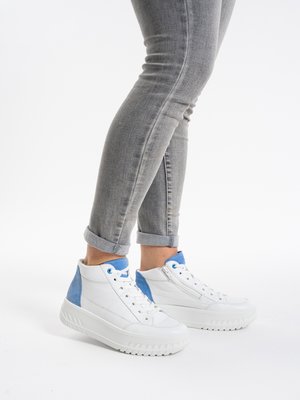 Person wearing white high-top sneakers with blue accents and gray rolled-up jeans, standing on white background.
