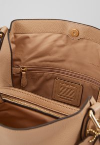 L'intérieur d'un sac à main en cuir tan présente une poche zippée, une texture lisse et une étiquette de marque. Doublé d'un tissu beige, fermé par un bouton-pression.