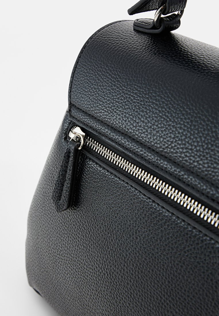 Sac à main en cuir noir avec texture granuleuse, sommet arrondi, fermeture éclair argentée et bandoulière amovible. La vue en gros plan met en valeur le détail de la fermeture éclair.