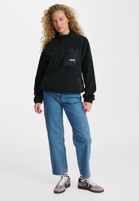 Sort fleecepuli med lynlås i halsen, der har en satinbrystlomme. Sammen med blå lige ben jeans og grå sneakers.