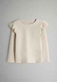 Ljus beige långärmad topp gjord av mjuk bomull, med volangdetaljer på axlarna och rund halsringning. Slät textur.