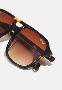 Gafas de sol de carey con lentes marrón degradado. Montura cuadrada, detalles metálicos y grabado "QUAY" en la patilla. Construcción de plástico durable.