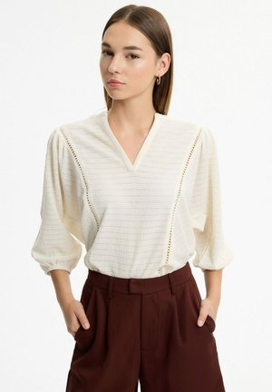 Mujer con pelo castaño liso lleva una blusa blanca con textura y mangas abullonadas de tres cuartos y pantalones marrones de talle alto con las manos en los bolsillos.