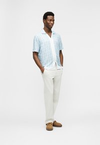 Άνδρας που στέκεται με τα χέρια στις τσέπες, φορώντας ανοιχτό μπλε πουκάμισο με μοτίβο και κοντά μανίκια, λευκό παντελόνι και καφέ παπούτσια σε λευκό φόντο.