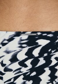 Plavati kostimi Calvin Klein se odlikujejo črno-belimi valovitimi vzorci z brušenim teksturo in so poudarjeni s pravokotno oznako na pasu.