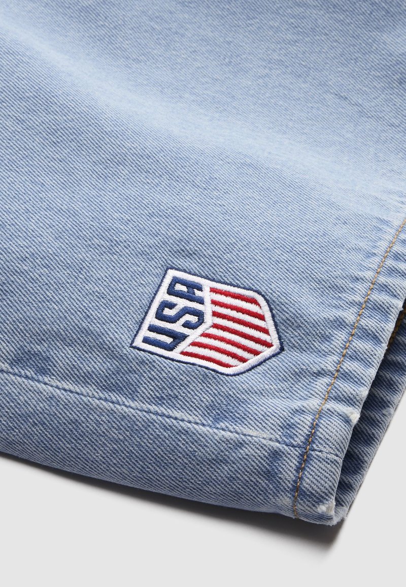 Gefalteter hellblauer Denimstoff mit einem aufgenähten USA-Emblem mit vertikalen roten Streifen und kräftigen blauen Buchstaben in der unteren Ecke.