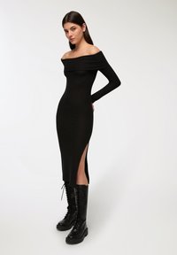 Abito nero a coste off-the-shoulder con maniche lunghe, fessure laterali e silhouette aderente, abbinato a stivali neri alti fino al ginocchio con lacci.