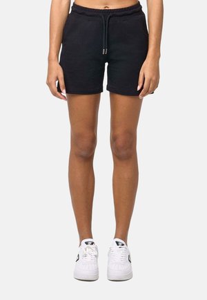 Femme portant un short noir à cordon et des baskets blanches, debout devant un fond clair uni, jambes et bas du torse visibles.
