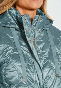 Gestepptes Puffermantel in Hellblau, mit hohem Kragen, Reißverschluss, Druckknöpfen und verstellbaren Trägern mit silbernen Akzenten.