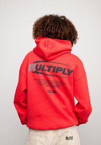 Felpa rossa con cappuccio e grande stampa nera "MULTIPLY" sulla schiena. Presenta dettagli testuali sotto il logo. Realizzata in tessuto morbido con una vestibilità rilassata.