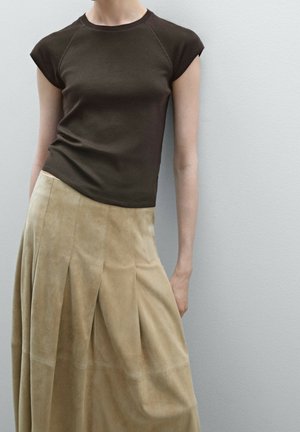 Personne portant un haut ajusté à manches courtes marron foncé et une jupe midi plissée beige, devant un mur gris clair.