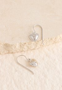 Silberne herzförmige Ohrringe mit polierter Oberfläche, die ein gebogenes Haken-Design und filigrane gezackte Details an den Rändern aufweisen.