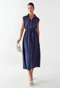 Abito senza maniche blu navy con bottone frontale, vita elasticizzata e due tasche sul petto. La modella tiene una borsa triangolare bianca.