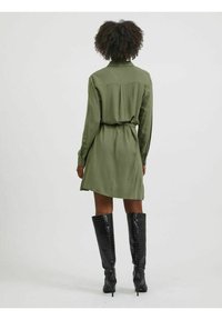 Olivgrön skjortklänning med långa ärmar, knäppmanschetter och ett bälte. Bärs med knähöga, svarta stövlar med krokodilprägling. Bakifrån.