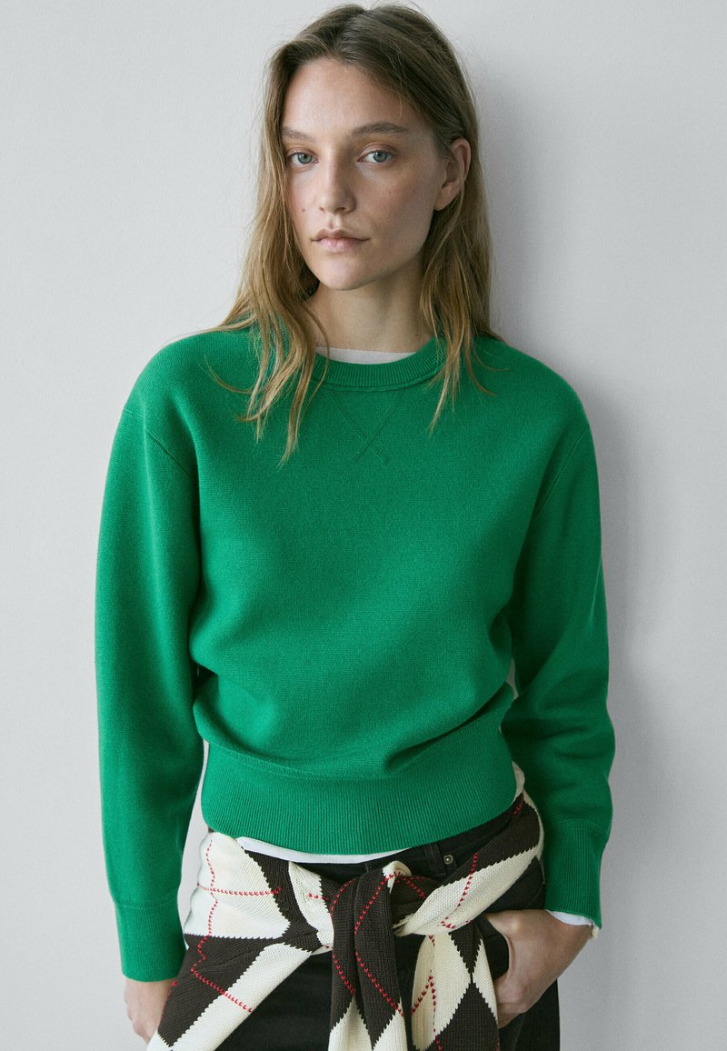 Ung kvinde med lyst brunt hår iført en grøn sweater og et sort-hvidternet stykke tøj bundet omkring taljen, stående op ad en væg.