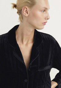 Camisa de pijama de terciopelo negro con cuello de muesca, textura de canalé vertical y cierre de botones. Cuenta con un bolsillo en el pecho.
