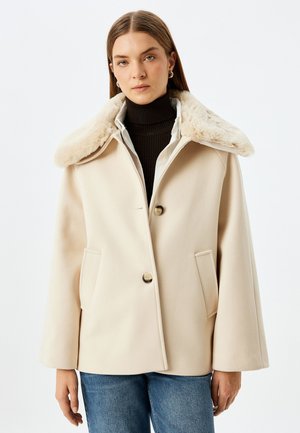 Beige uldblandet frakke med en stor faux fur krave, to frontlommer og en enkelt knaplukning. Lange, let udvidede ærmer.
