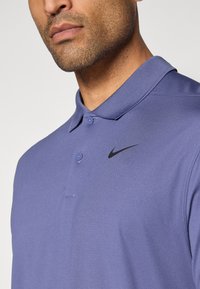 Fialová polo košeľa vyrobená z textúrovaného materiálu, s krátkym golierom, dvoma gombíkmi a čiernym logom Nike na ľavom prsnom vrecku.