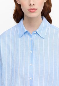 Light blue button-up shirt with white vertical stripes, featuring a pointed collar and three buttons visible at the front.