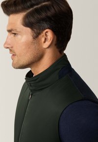 Gilet verde scuro con colletto alto, caratterizzato da una zip e dettagli blu navy. Texture liscia e design aderente, che mette in evidenza un profilo laterale.