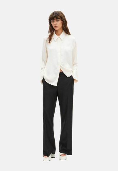 Koszula z długim rękawem w kolorze jasnego kremu, zapinana na guziki, z ostrym kołnierzem, zestawiona z czarnymi spodnimi o szerokich nogawkach oraz masywnymi białymi sandałami na platformie.