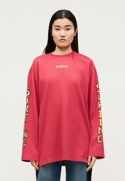 Γυναίκα που φοράει άνετο κόκκινο μακρυμάνικο πουκάμισο με το «STIEGLITZ» στο στήθος και το «SURFING» τυπωμένο κάθετα και στα δύο μανίκια, σε συνδυασμό με τζιν.