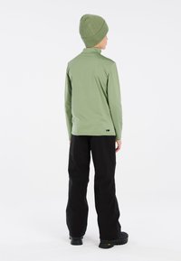 Maglia verde a maniche lunghe con colletto alto, abbinata a pantaloni neri e a un berretto verde a coste coordinato. Tessuto liscio e design aderente.