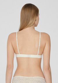 Světle béžová podprsenka s nastavitelnými ramínky, zapínáním na háčky a hladkou texturou. Minimální design zahrnuje jednoduchá záda bez ozdob.