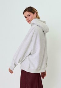 Gaišpelēka pārmērīga sudraba pelēka kapuce ar lielu kapuci, pazeminātiem pleciem un ribotām aprocēm. Valkāta ar bordo svārkiem uz gaiša fona.