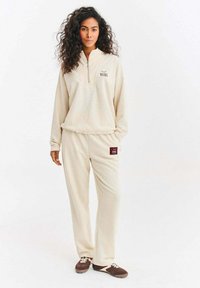 Béžový fleecový pulovr se zipovým límečkem a vyšitým logem, doplněný o odpovídající fleecové kalhoty s náprsným kapsou a stahovací šňůrkou v pase.