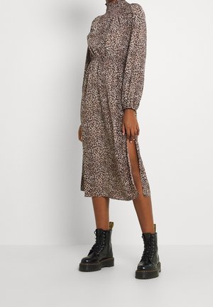 Persoon die een bruine midi-jurk met luipaardprint draagt, lange mouwen en een hoge split, gecombineerd met zwarte veterplateauboots.