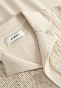 Krémovo sfarbený pletený sveter s golierom, gombíkom a textúrovaným otvoreným vzorom pletenia, s menovkou značky "GABBA" vo vnútri goliera.