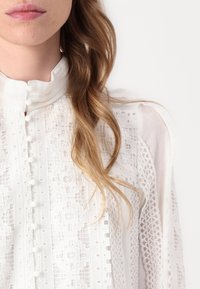 Hvit bluse med teksturert stoff, detaljert broderi, høy krage og knappelukking foran. Lange, gjennomsiktige ermer med blondekant.