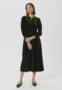 Mørk grønn velurkjole med smock-bodice, lange puffede ermer og volangmansjetter. Midtlengde design som faller under kneet. Svart flate sko.