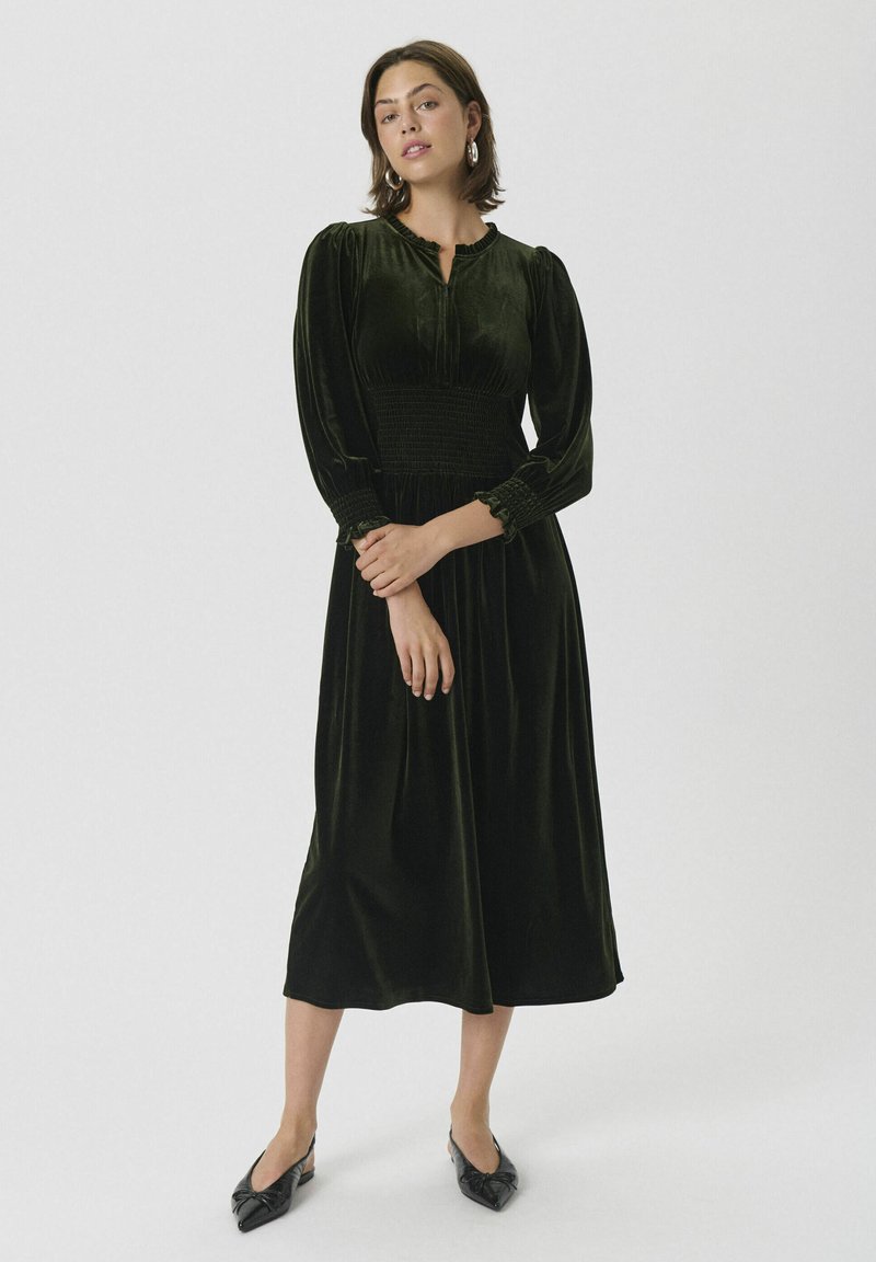 Mørk grønn velurkjole med smock-bodice, lange puffede ermer og volangmansjetter. Midtlengde design som faller under kneet. Svart flate sko.