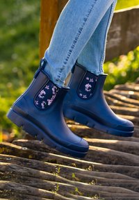 Dunkelblaue Gummistiefel bis zur Knöchel mit floralen Stoffakzenten, strukturierten Sohlen und Zuglaschen, getragen mit hellblauen Jeans auf einer Holzoberfläche.