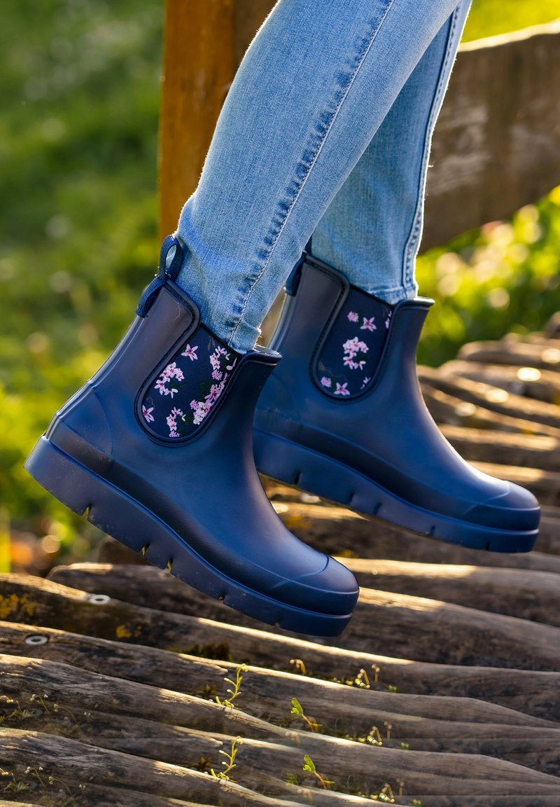 Dunkelblaue Gummistiefel bis zur Knöchel mit floralen Stoffakzenten, strukturierten Sohlen und Zuglaschen, getragen mit hellblauen Jeans auf einer Holzoberfläche.