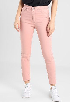 Kobieta w różowych, obcisłych dżinsach z wysokim stanem, czarnym włożonym do środka topie i białych tenisówkach na sznurowadła, stojąca na jednolitym jasnoszarym tle.