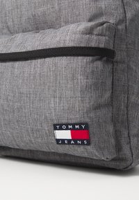 Šedý látkový batoh s texturovaným povrchem, který má přední kapsu, černý zip a obdélníkovou nášivku Tommy Jeans v červené, bílé a modré barvě.