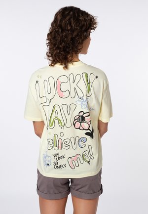 Person with curly hair wearing light yellow t-shirt with colorful text "LUCKY DAY believe me! You look so lovely" and flower drawings on back.