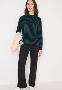 Maglietta a maniche lunghe a righe in verde scuro e nero, con un cappuccio staffa. Abbinata a jeans neri a zampa d'elefante e una giacca beige chiaro. Sneakers bianche.