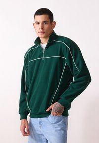 Grøn fleece halv-zip sweatshirt med hvide detaljer. Lange ærmer og en afslappet pasform; har en høj krave og en lynlås ved halsen.