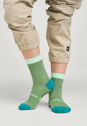 Persona che indossa pantaloni beige con risvolto e calzini verdi con punte e tacchi turchesi, in piedi su una superficie grigia.