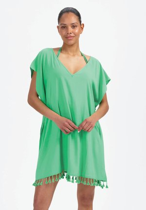 Donna che indossa un abito copricostume verde con scollo a V ampio e nappine all'orlo, in piedi contro uno sfondo bianco semplice.